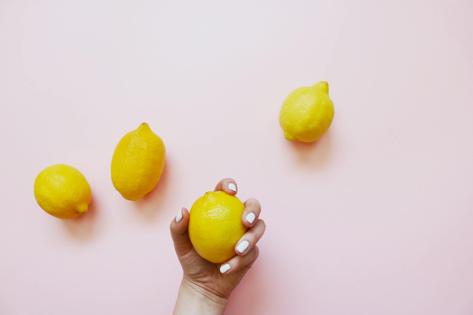 A hand holding a lemon on a soft pink background with additional lemons nearby, symbolizing stress relief and self-care
