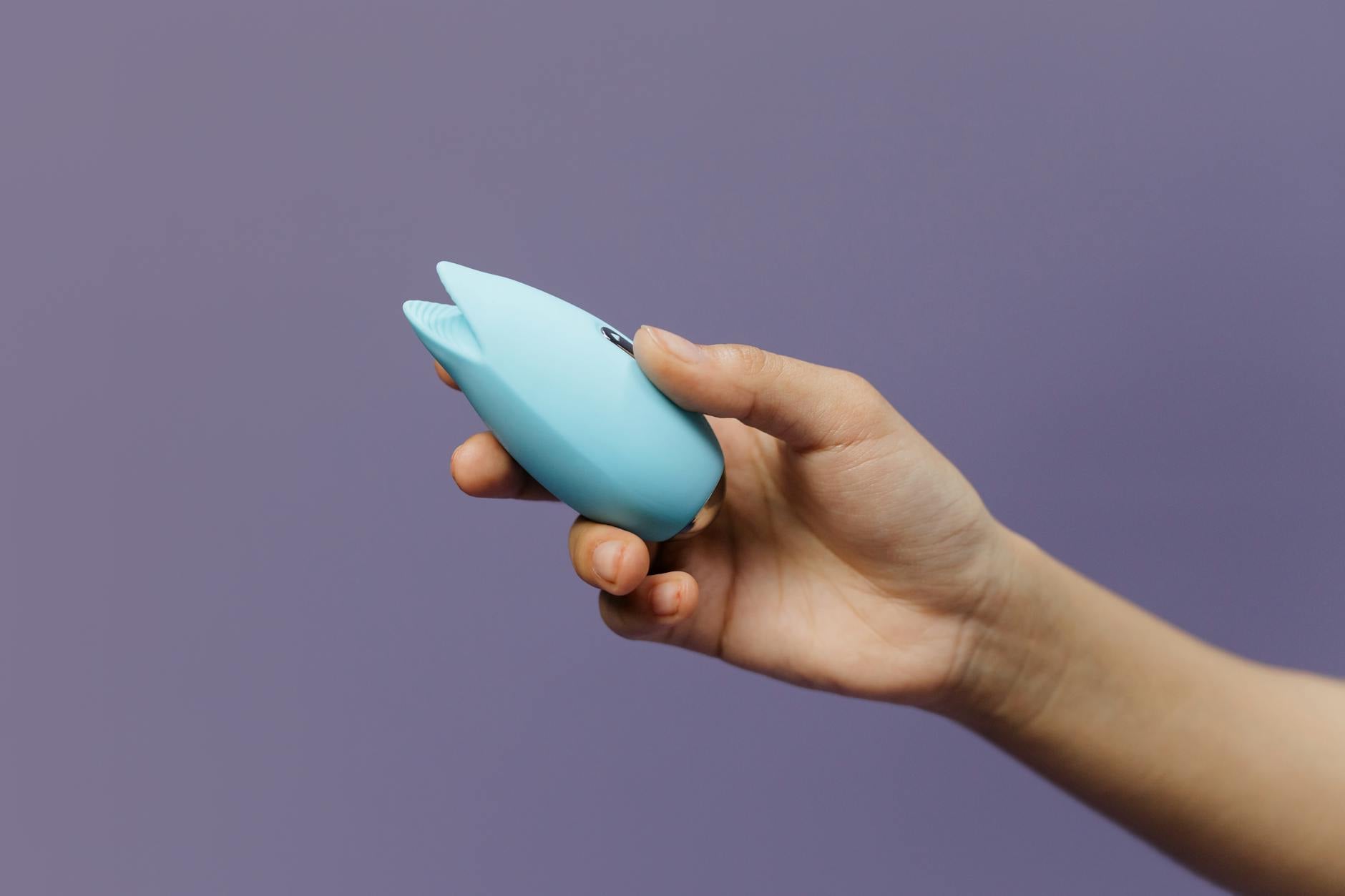 A blue silicone clitoral vibrator held in hand against a purple background, representing gentle pleasure and self-care.
